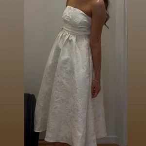 Vit axelbandslös klänning med strukturmönster - Säljer en elegant vit axelbandslös klänning med vackert strukturmönster och markerad midja. Klänningen har en utsvängd kjol och knytning i ryggen för extra passform. Perfekt för speciella tillfällen. Använd en kväll och kemtvättad. Passar XS-S