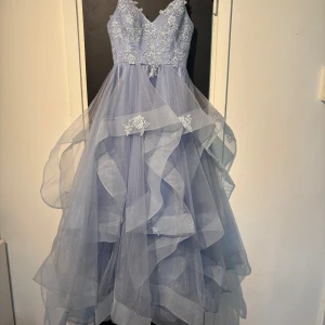 Ljusblå Bal/Festklänning - Drömmig ljusblå långklänning med v-ringad överdel, tunna axelband och vackra spetsdetaljer. Kjolen är i flera lager tyll med volanger och broderade blommor. Perfekt för bal eller fest. Ger en riktigt sagolik känsla!