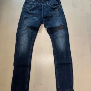 Mörkblå jeans med slitningar - Säljer ett par mörkblå jeans med snygga slitningar och klassisk femficksdesign. Jeansen har normal passform och raka ben, perfekt till vardags. Stängs med dragkedja och knapp framtill.