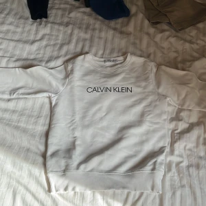 Vit sweatshirt från Calvin Klein Jeans - Säljer en stilren vit sweatshirt från Calvin Klein Jeans med svart logotyp på bröstet och en liten 'JEANS'-patch bak. Tröjan har rund halsringning och långa ärmar. Perfekt för en clean och enkel look.