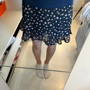 Blommig volangkjol - Supersöt svart kjol från SHEIN med små rosa och vita blommor. Kjolen har volang nertill och en diskret dragkedja i sidan. Perfekt för dig som gillar en feminin och lekfull stil. Super svalt och skönt material. Kjolen är i storlek XS. 