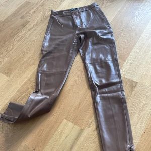 Bruna skinnbyxor från Zara - Snygga bruna skinnbyxor från Zara med hög midja och raka ben. Byxorna har dragkedja och knapp framtill samt diskreta fickor. Perfekta för dig som vill ha en trendig och stilren look.