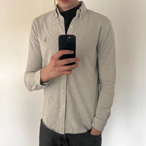 Ljusgrå skjorta från Ralph Lauren - Säljer en stilren ljusgrå skjorta från Ralph Lauren med klassisk button-down krage och broderad logga på bröstet. Skjortan har långa ärmar och knäppning framtill. Perfekt för dig som gillar en clean och tidlös look.