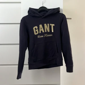 Mörkblå GANT hoodie - Säljer en mörkblå hoodie från GANT med guldigt broderat tryck 'GANT New Haven' på bröstet. Tröjan har huva och en stor magficka framtill. Perfekt för dig som gillar klassisk och sportig stil. Inga defekter. 💗💗