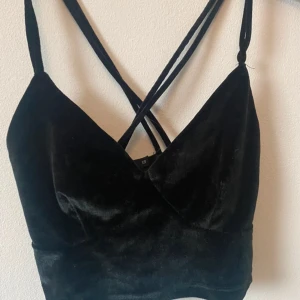 Svart sammetstopp med smala axelband från BikBok - Säljer en svart croppad sammetstopp från BikBok med smala korsade axelband. Toppen har en omlottliknande v-ringning och är perfekt för att skapa en trendig look.Frakt tillkommer