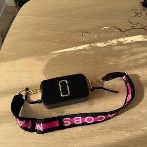 Svart snapshot väska från Marc Jacobs - Säljer en svart axelväska från Marc Jacobs med bred axelrem i svart, rosa och vitt med logga. Väskan har guldfärgade detaljer och dubbla dragkedjor upptill samt en vit panel på ovansidan. Perfekt storlek för det viktigaste.