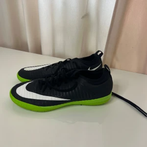 Nike Mercurial futsal skor - Säljer ett par Nike Mercurial futsalskor i svart med vit swoosh och grön sula. Skorna har snörning och är designade för inomhusspel. Perfekta för dig som vill ha snabba och lätta skor på planen.