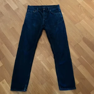 Mörkblå jeans från Nudie Jeans - Säljer ett par klassiska mörkblå jeans från Nudie Jeans med raka ben och femficksmodell. Byxorna har tydliga Nudie-detaljer på bakfickorna och läderpatch i midjan. Perfekta för dig som gillar stilrena och tidlösa jeans.