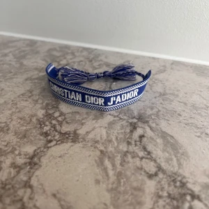 Blått armband från Christian Dior J'ADIOR - Säljer ett blått tygarmband från Christian Dior med texten 'J'ADIOR' broderad i vitt. Armbandet har justerbar knytning med tofsar i ändarna och är en snygg accessoar med exklusiv känsla.