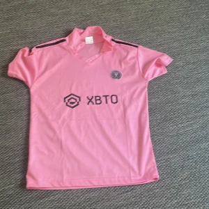 Inter Miami CF rosa fotbollströja - Säljer en rosa Inter Miami CF fotbollströja med svarta detaljer och XBTO sponsortryck på bröstet. Tröjan har korta ärmar, klassisk krage och klubbmärke på vänster bröst. Perfekt för dig som vill sticka ut på planen eller läktaren.