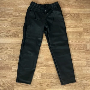 Byxor i skinnimitation från Carin Wester - Snygga svarta byxor från Carin Wester i skinnimitation med resår och snörning i midjan. Byxorna har raka ben och en stilren look som passar till många olika outfits.