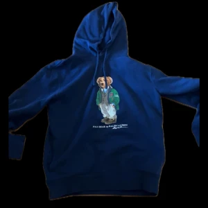 Blå hoodie med Polo Bear från Ralph Lauren - Säljer en blå hoodie från Ralph Lauren med den klassiska Polo Bear-trycket på bröstet. Tröjan har huva med dragsko och en avslappnad passform. Perfekt för dig som gillar streetwear och ikoniska motiv.