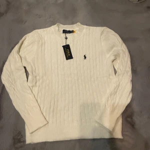Vit kabelstickad tröja från Polo Ralph Lauren - Säljer en klassisk vit kabelstickad tröja från Polo Ralph Lauren med v-ringning och broderad logga på bröstet. Tröjan har lång ärm och är tillverkad i mjukt material som passar perfekt till många olika outfits.