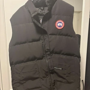 Svart dunväst från Canada Goose - Säljer en svart dunväst från Canada Goose i modellen Fusion XL. Västen har två stora fickor framtill, dragkedja och klassisk Canada Goose-logga på bröstet. Perfekt för kyliga dagar och har en stilren, normal passform.