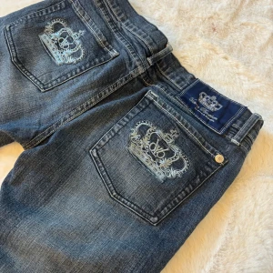 Mörkblå jeans med broderad krona från Victoria Beckham - Snygga mörkblå jeans från Victoria Beckham med broderad krona på bakfickorna och patch med logga. Klassisk femficksmodell i jeansmaterial med raka ben och normal passform.