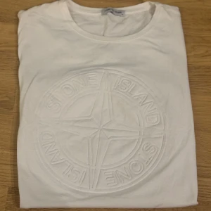 Vit t-shirt från Stone Island - Vit t-shirt från Stone Island med stort broderat logotypmönster framtill. Klassisk rund halsringning och enkel, stilren design som passar till det mesta.