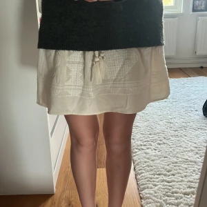 Vit kortkjol med tofsar från Zara - Supersöt vit kortkjol från Zara med smock i midjan och dekorativa tofsar framtill. Kjolen har ett diskret mönster i tyget och knappar längs framsidan. Perfekt för en fräsch och somrig look! Aldrig använd 