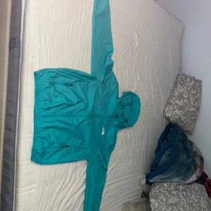 Turkos vindjacka från The North Face - Säljer en turkos vindjacka från The North Face med huva och dragkedja. Jackan har två sidofickor med dragkedja och är perfekt för blåsiga dagar. Enkel och sportig design med logga på bröstet.