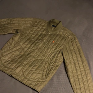 Olivgrön quiltad jacka från Polo Ralph Lauren - Säljer en olivgrön quiltad jacka från Polo Ralph Lauren med orange broderad logga på bröstet. Säljer för att den ej passar längre. Hör av dig vid minsta fundering!
