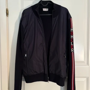 Svart Moncler jacka med logga på ärmen - Säljer en svart jacka från Moncler med dragkedja och ribbade muddar. Jackan har lång ärm med Moncler-logga i vitt och rött längs ena ärmen samt diskreta fickor med dragkedja. Snygg och sportig design med klassiska Moncler-detaljer. Ny pris 920 euro