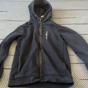 Svart hoodie från Sail Racing - Mörkgrå hoodie från Sail Racing med dragkedja framtill och huva. Tröjan har en bröstficka med dragkedja och logga på bröstet. Perfekt för kyliga dagar och enkel att matcha med det mesta.