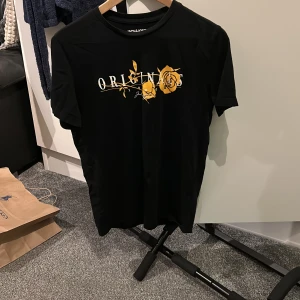 Svart t-shirt med gul ros från Jack & Jones - Säljer en svart t-shirt från Jack & Jones med tryck av texten 'ORIGINS' och en gul ros på bröstet. T-shirten har rund halsringning och korta ärmar. Perfekt för en stilren och trendig look.