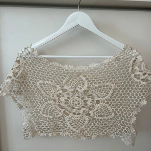 Beige virkad topp - Säljer en beige virkad topp med ett stort blommönster framtill. Toppen är kort och har ett luftigt, virkat utseende som ger en bohemisk känsla. Står ingen storlek, men skulle uppskatta att det är XS. 