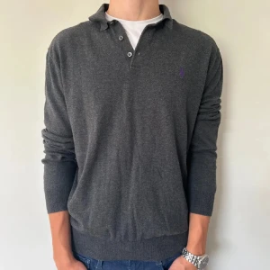  pikétröja från Polo Ralph Lauren - Säljer en klassisk mörkgrå pikétröja från Polo Ralph Lauren i mjuk pima-bomull. Tröjan har långa ärmar, krage och knappar framtill samt en lila broderad logga på bröstet. Perfekt för en stilren och avslappnad look.