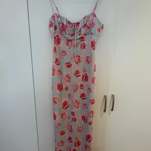 Långklänning med rosor och smala axelband - Säljer en långklänning med smala axelband och blommigt mönster i rött och rosa på ljus bakgrund. Klänningen har en feminin passform och knytning framtill vid bysten. Perfekt för sommarens alla tillfällen!