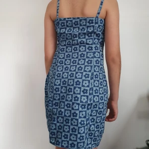 Blå blommig kortklänning med smala axelband - Säljer en blå kortklänning med smala axelband och blommigt rutmönster. Klänningen har figurnära passform och är tillverkad i ett mjukt jeanstyg. Perfekt för sommarens varma dagar!