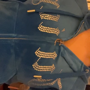 Blå hoodie från Juicy Couture - Lyxigt mjuk hoodie med dragkedja och huva från Juicy Couture. Klassisk passform och ikonisk design med stora bokstäver i glittrande strass framtill. Perfekt för en avslappnad men stilren look. Endast använd en gång.