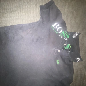 Svart t-shirt från Hugo Boss med tryck - Svart t-shirt från Hugo Boss med stort grönt och vitt BOSS-tryck på bröstet och detaljer på kragen. Klassisk passform med korta ärmar och knappslå vid halsen. Perfekt för en stilren och sportig look.