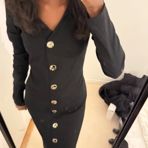 Svart ribbad långklänning med guldknappar - Säljer en svart ribbad långklänning med v-ringning och långa ärmar. Klänningen har stora dekorativa guldknappar längs hela framsidan och en figurnära passform. Perfekt för dig som gillar stilrena och eleganta plagg.