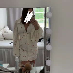 Vit blommig kortklänning med v-ringning och puffärmar från adoore - Superfin vit kortklänning med små röda blommor, djup v-ringning och långa puffiga ärmar från adooore Klänningen har knappar framtill och markerad midja som ger en snygg siluett. Storlek 38🩷 Endast använd en gång