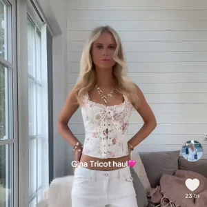 Blommig korsettopp med spets från Gina Tricot - Superfin blommig korsettopp från Gina Tricot i ljus cremefärg med rosa och lila blommor. Toppen har spetsdetaljer vid kanterna och snörning framtill. Perfekt för en trendig och romantisk look.