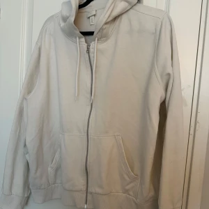 Vit hoodie med dragkedja från H&M - Säljer en enkel och stilren vit hoodie från H&M Basics i storlek XL. Tröjan har dragkedja framtill, huva med dragsko och två stora fickor. Tillverkad i bomull och polyester för en mjuk och bekväm känsla.