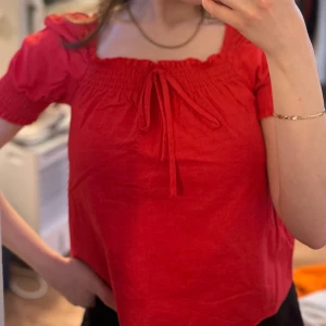 Röd blus med knyt och puffärmar - Säljer en söt röd blus med smock upptill, puffiga korta ärmar och knyt framtill. Perfekt för dig som gillar färg och vill ha en feminin look. Blusen har en fyrkantig ringning och är tillverkad i ett mjukt bomullstyg.