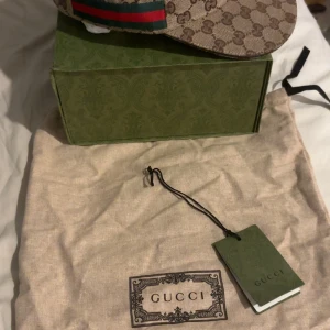 Beige keps från Gucci med GG-mönster - Snygg beige keps från Gucci med klassiskt GG-monogram över hela kepsen och grönt/rött band på sidan. Kepsen har böjd skärm och justerbar rem bak. Perfekt för dig som gillar exklusiva accessoarer.
