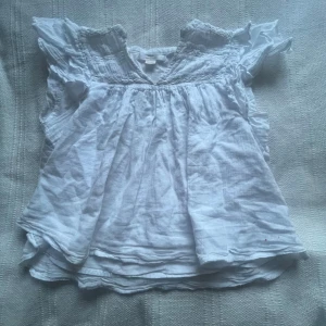 Vit blus med volang från Gina Tricot - Söt vit blus från Gina Tricot med volangdetaljer vid axlarna och lätt rynk vid halsringningen. Blusen är ärmlös och har en luftig passform, perfekt för varma dagar. Tillverkad i ett tunt och mjukt material.