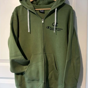 Grön hoodie med dragkedja från Champion - Säljer en grön hoodie från Champion med dragkedja framtill och huva med vita snören. Väldigt skön insida. Aldrig använd