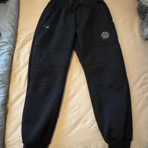 Svarta mjukisbyxor med ribbade detaljer - Svarta mjukisbyxor från Fashion med ribbade partier över knäna och dragsko i midjan. Byxorna har resår vid bensluten och en liten logga på ena benet. Tillverkade i bomull och polyester för en mjuk känsla.