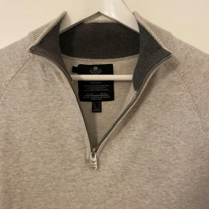 Ljusgrå halvzip-tröja från Threadbare - Säljer en stilren ljusgrå tröja från Threadbare med halv dragkedja och hög krage. Tröjan har lång ärm och ribbade detaljer vid krage och ärmslut. Perfekt för lager-på-lager och enkel att matcha.