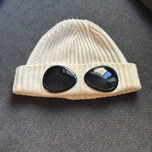 Vit stickad mössa från C.P. Company - Vit ribbstickad mössa från C.P. Company med unika svarta glasögonlinser framtill. Klassisk design med uppvikt kant och tydlig logga på insidan. Skick 9/10 använd ett fåtal gånger 