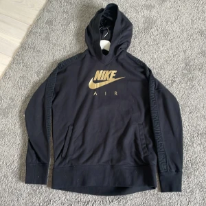 Svart hoodie från Nike Air med guldtryck - Svart hoodie från Nike Air med stort guldigt logotryck på bröstet. Tröjan har huva och långa ärmar med diskret mönster längs ärmarna. Perfekt för en sportig och avslappnad stil.