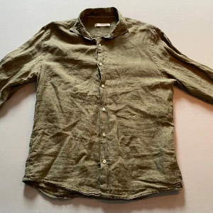 Olivgrön linneskjorta från MNG - Säljer en olivgrön skjorta i linne från MNG (MANGO). Skjortan har klassisk krage, långa ärmar och knäppning framtill med vita knappar. Perfekt för en avslappnad och stilren look. Storlek M.