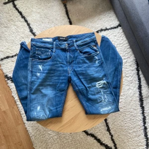 Blå slitna Replay jeans - Hej👋 Säljer ett par blå jeans från Replay med slitningar och lätt tvättad look👖 Klassisk femficksmodell med knappgylf och läderpatch bak i midjan. Jeansen har raka ben och normal passform🧷 Jeansen har Anbass modell!  Storleken på jeansen är W33 L34📐 Kom privat för mått📏  Priset är INTE ristat i sten så kom gärna med prisförslag💵  Vid frågor: Meddela oss!📩.