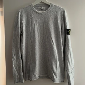 Grå långärmad tröja från Stone Island - Säljer en stilren grå långärmad tröja från Stone Island med klassisk patch på vänster ärm. Tröjan har rund halsringning och är tillverkad i mjuk bomull, perfekt för en clean och avslappnad look.