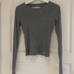 Grå långärmad tröja från Hollister - Säljer en enkel och stilren grå långärmad tröja från Hollister. Tröjan är i mjukt material och har en tight passform, perfekt att bära till jeans eller kjol. Liten Hollister-logga nertill på framsidan.