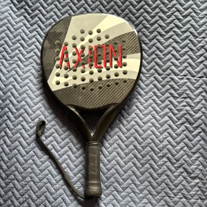 Padelracket AXION i kolfiber - Säljer ett padelracket från AXION i svart och grått med vita detaljer. Racketen är tillverkad i kolfiber och har ett handtag med handledsrem. Perfekt för dig som spelar padel!