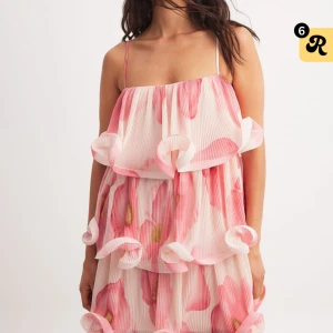 Rosa och vit volangklänning med blommigt mönster - Superfin klänning med tunna axelband och flera lager volanger. Klänningen har ett blommigt mönster i rosa och vitt, och är perfekt för sommarens alla tillfällen. Lätt och luftig modell. Helt ny i plast med lappen kvar.